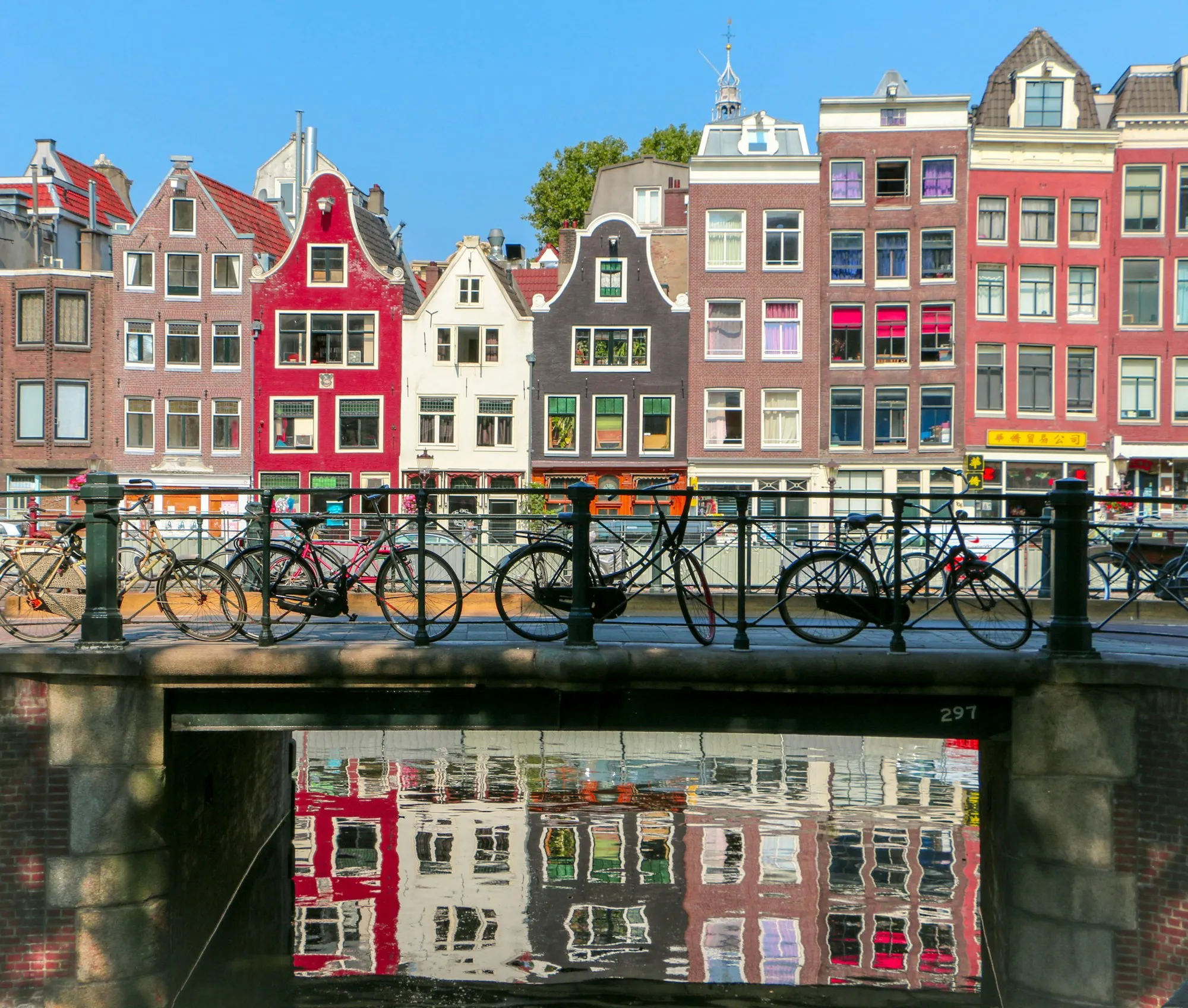 Colourful gabled canal houses behind a bridge railing lined with parked bicycles, reflected in the water below