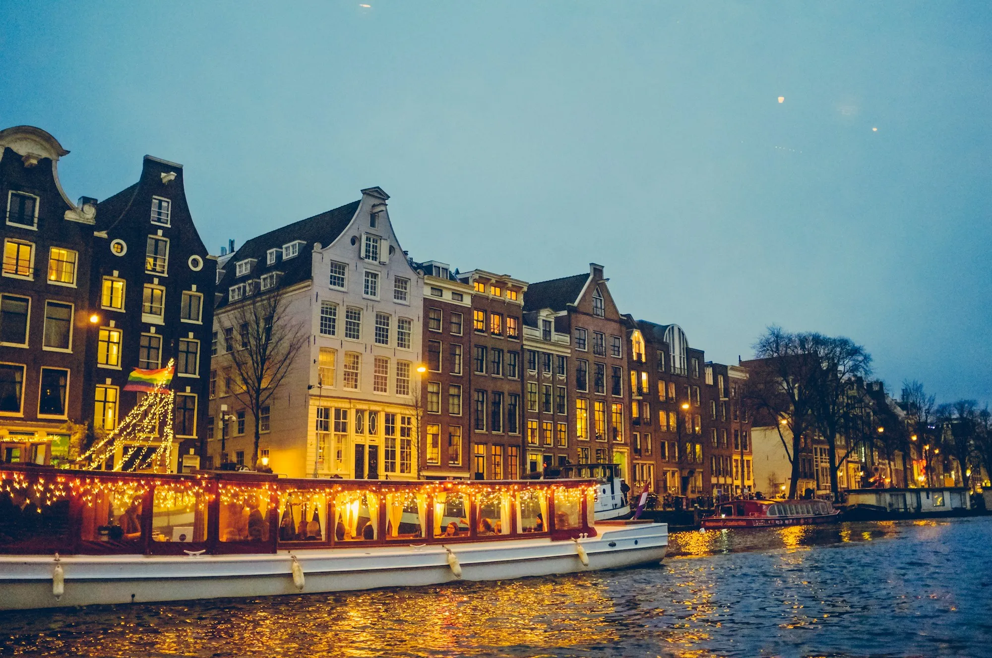 An Amsterdam canal at dusk — gabled houses and a lantern-lit boat glowing warm against a blue evening sky
