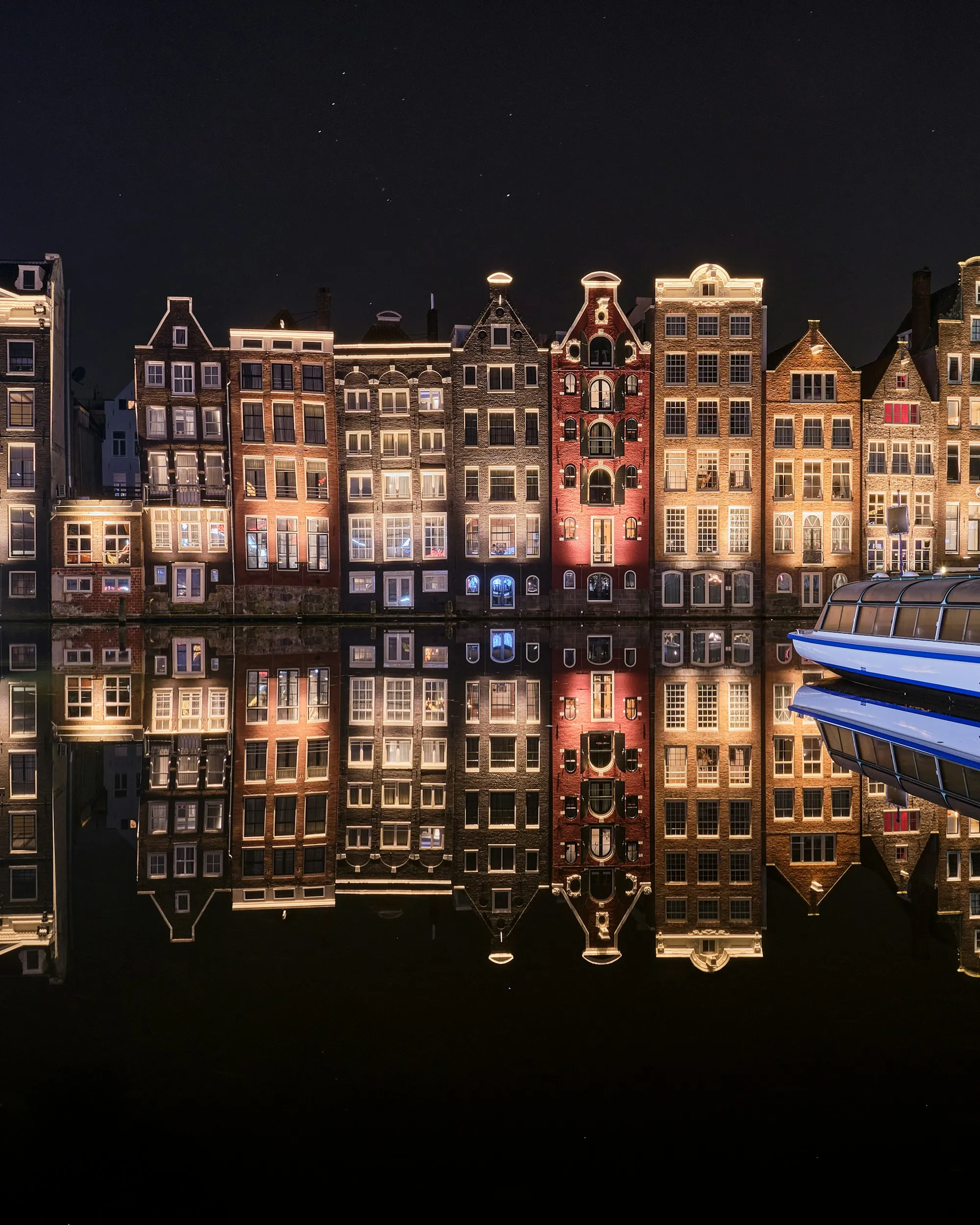 A row of Amsterdam canal houses lit up at night, mirror-reflected in the still black water, a glass tour boat entering from the right