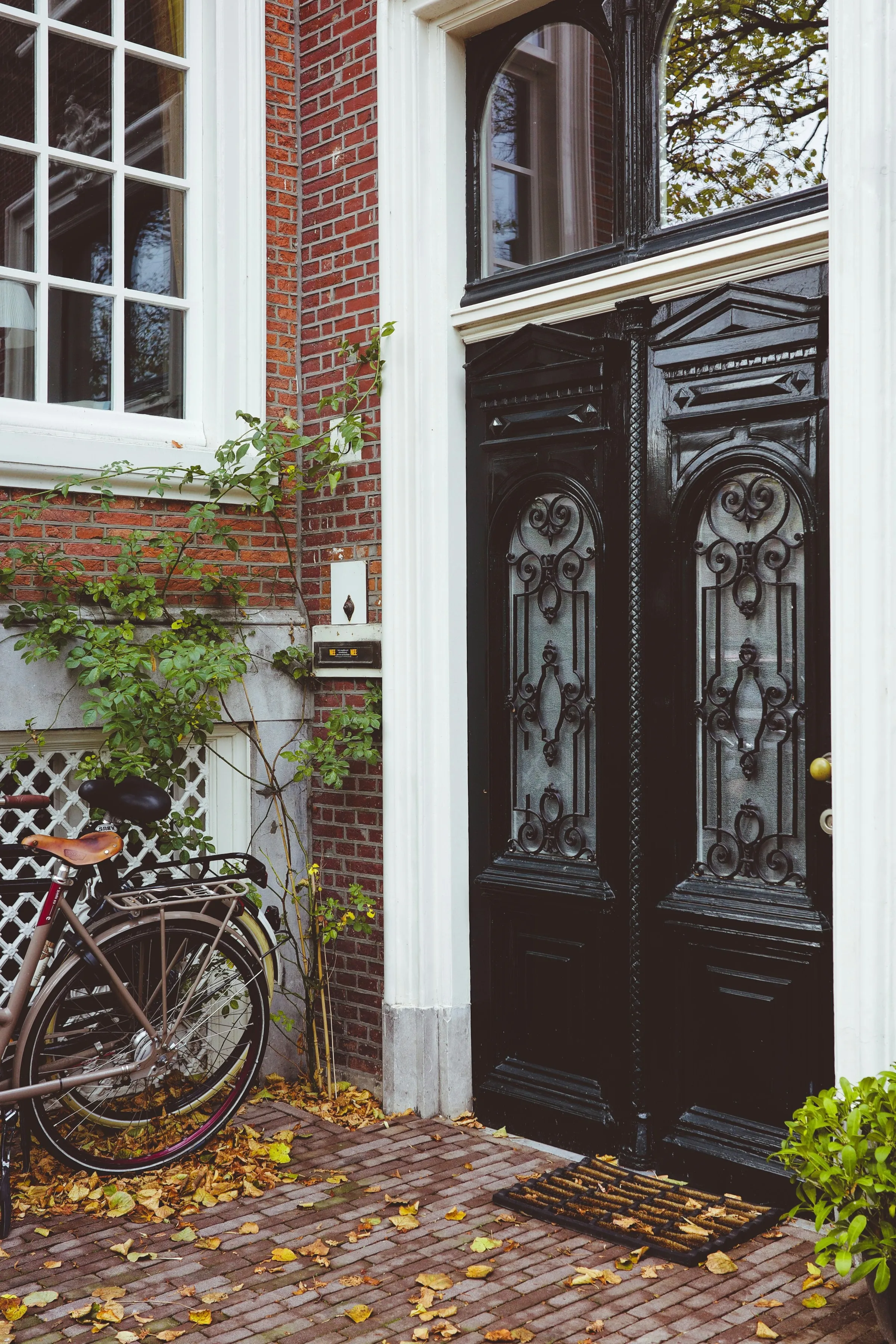 A bicycle parked against a brick wall beside a tall dark double door, autumn leaves on the pavement